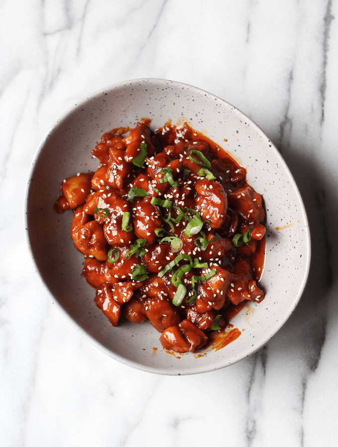 Red gochujang chicken in white bowl topped with spring onions.