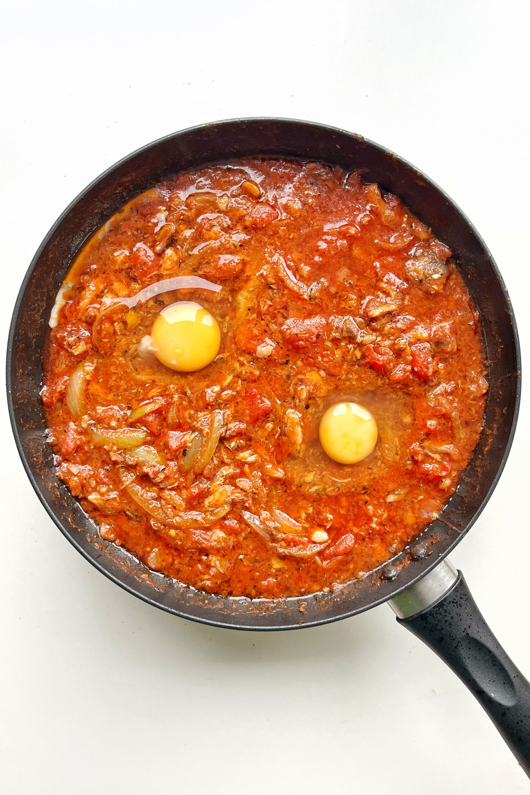 Black pan with tomato and sardine sauce with two eggs cracked into the middle.
