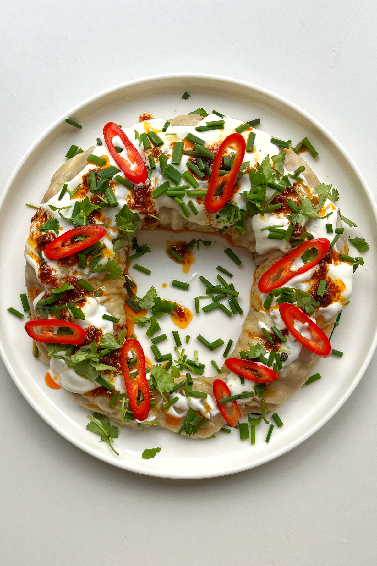 Dumpling wreath on a white plate topped with yoghurt, harissa oil, chives, and sliced chilli. 