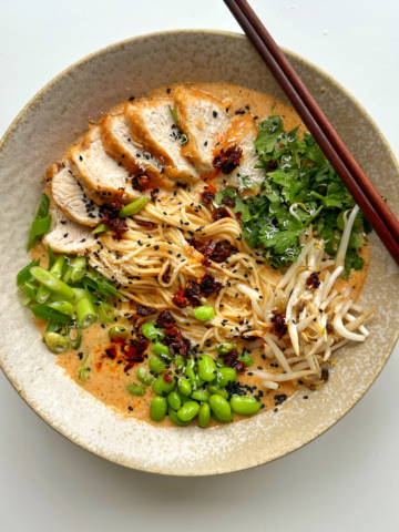 Marry me chicken ramen in a ramen bowl with chopsticks topped with chicken, edamame beans, coriander, beansprouts, and spring onions.