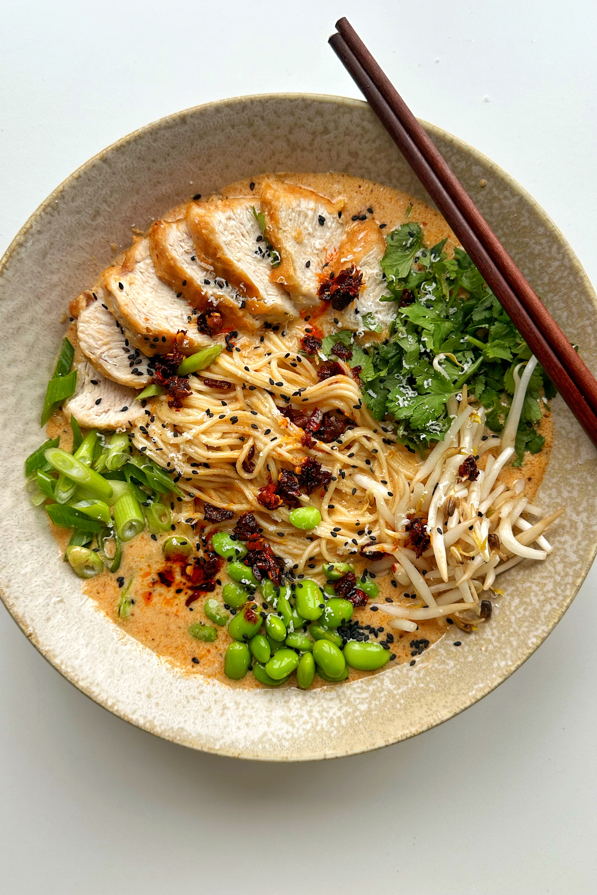 Marry me chicken ramen in a ramen bowl with chopsticks topped with chicken, edamame beans, coriander, beansprouts, and spring onions. 