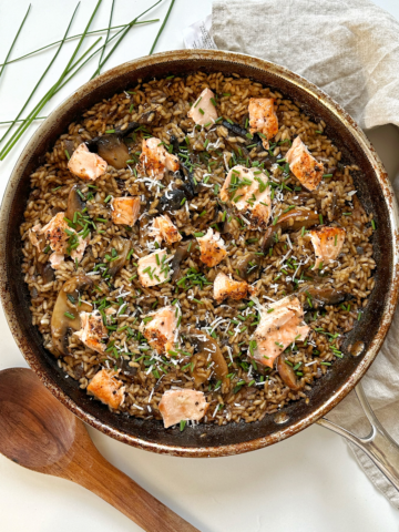 Salmon and mushroom risotto in frying pan with wooden spoon, chives, and napkin surrounding it.