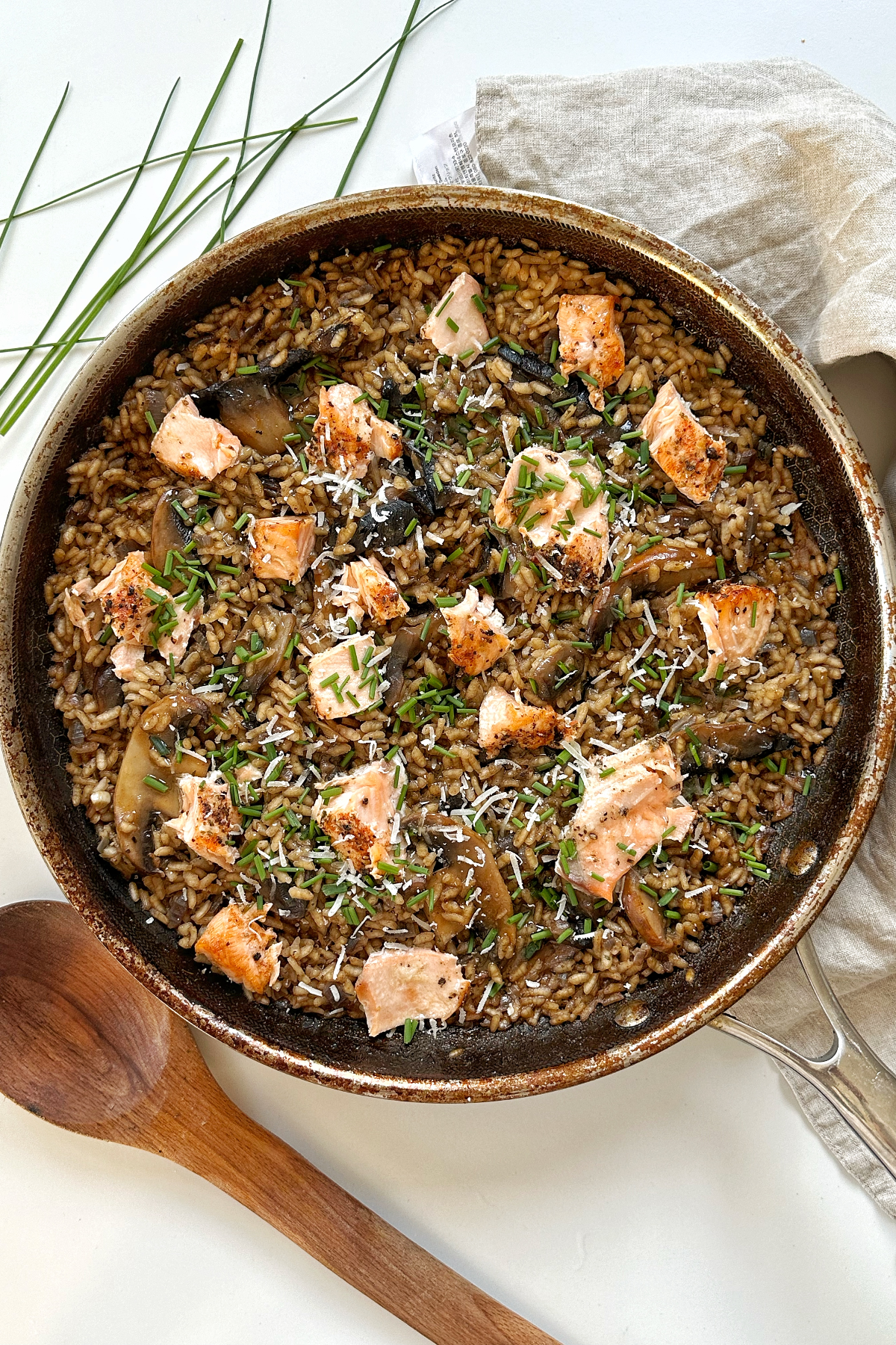 Salmon and mushroom risotto in frying pan with wooden spoon, chives, and napkin surrounding it. 