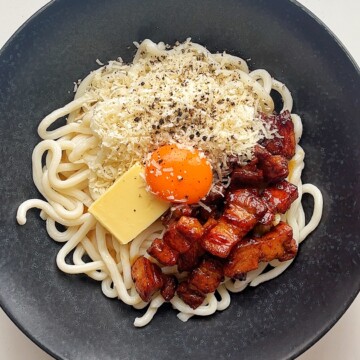 Black bowl with udon noodles, fried pork belly, grated parmesan, knob of butter, and egg yolk.