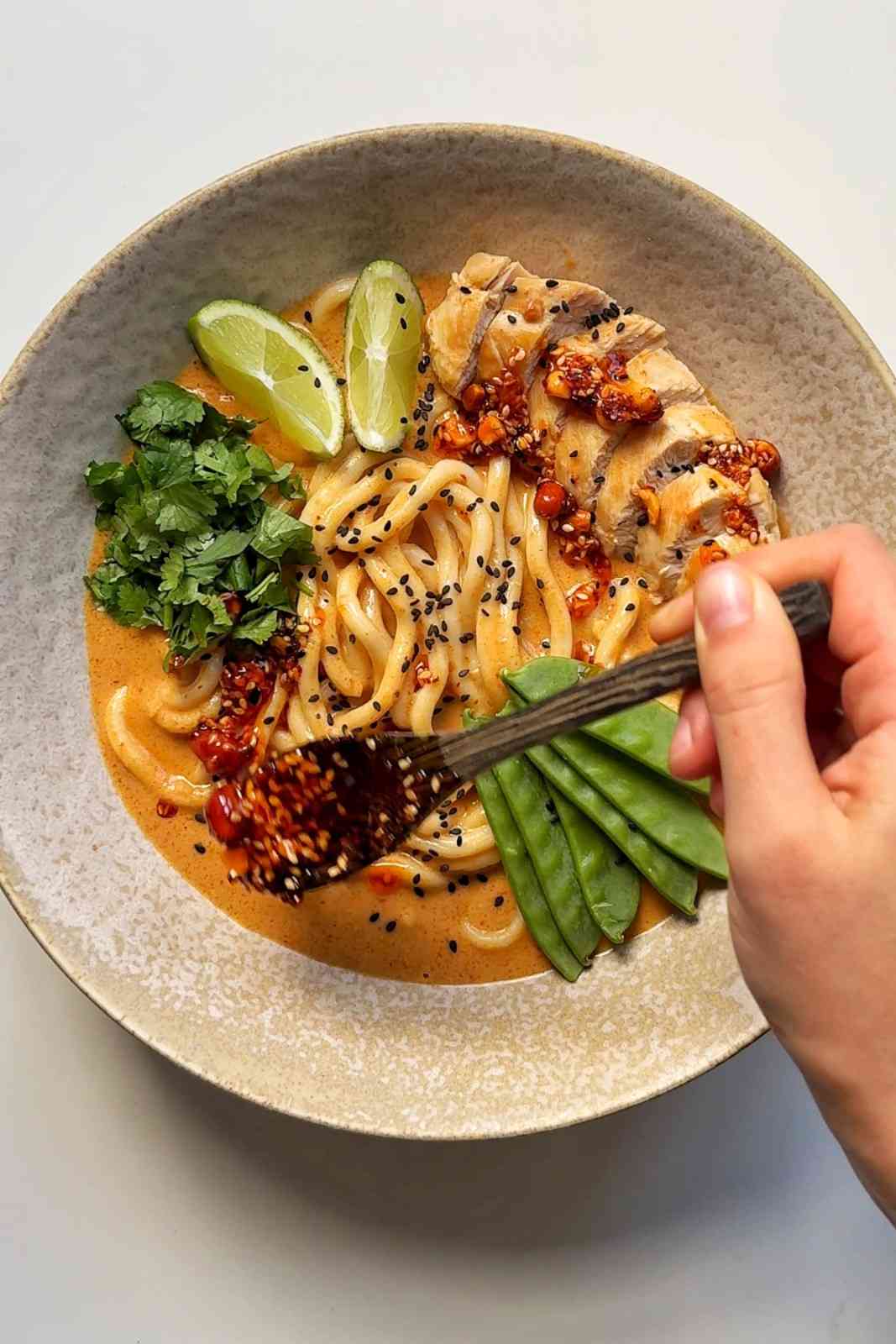 A bowl of creamy peanut butter satay ramen topped with sliced chicken, snow peas, fresh cilantro, and lime wedges, with a hand drizzling a spicy chili peanut sauce over the noodles.