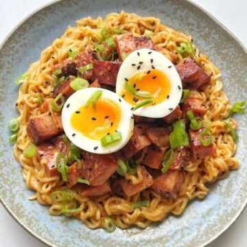 A bowl of instant ramen noodles topped with caramelized cubes of fried Spam, a halved soft-boiled egg, and sliced green onions, showcasing the viral Spam hack.
