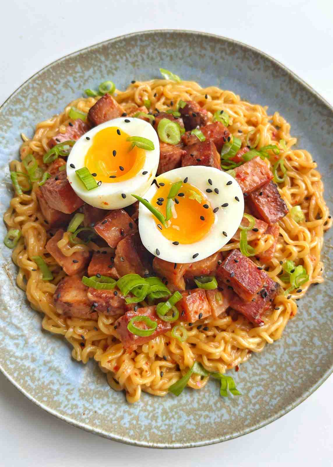 A close-up, top-down view of a bowl of instant ramen topped with cubes of pan-fried, caramelized Spam, two jammy soft-boiled egg halves, fresh green spring onions, and a sprinkle of black sesame seeds.