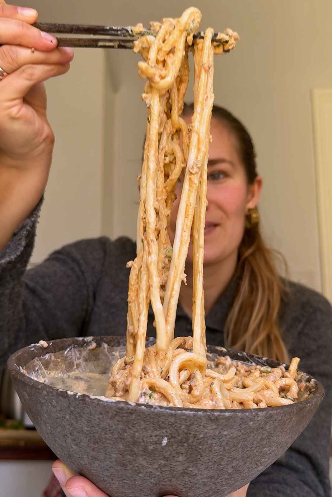 Emily Roz lifting long creamy mapo tofu udon noodles from a stone bowl with chopsticks, smiling at the camera