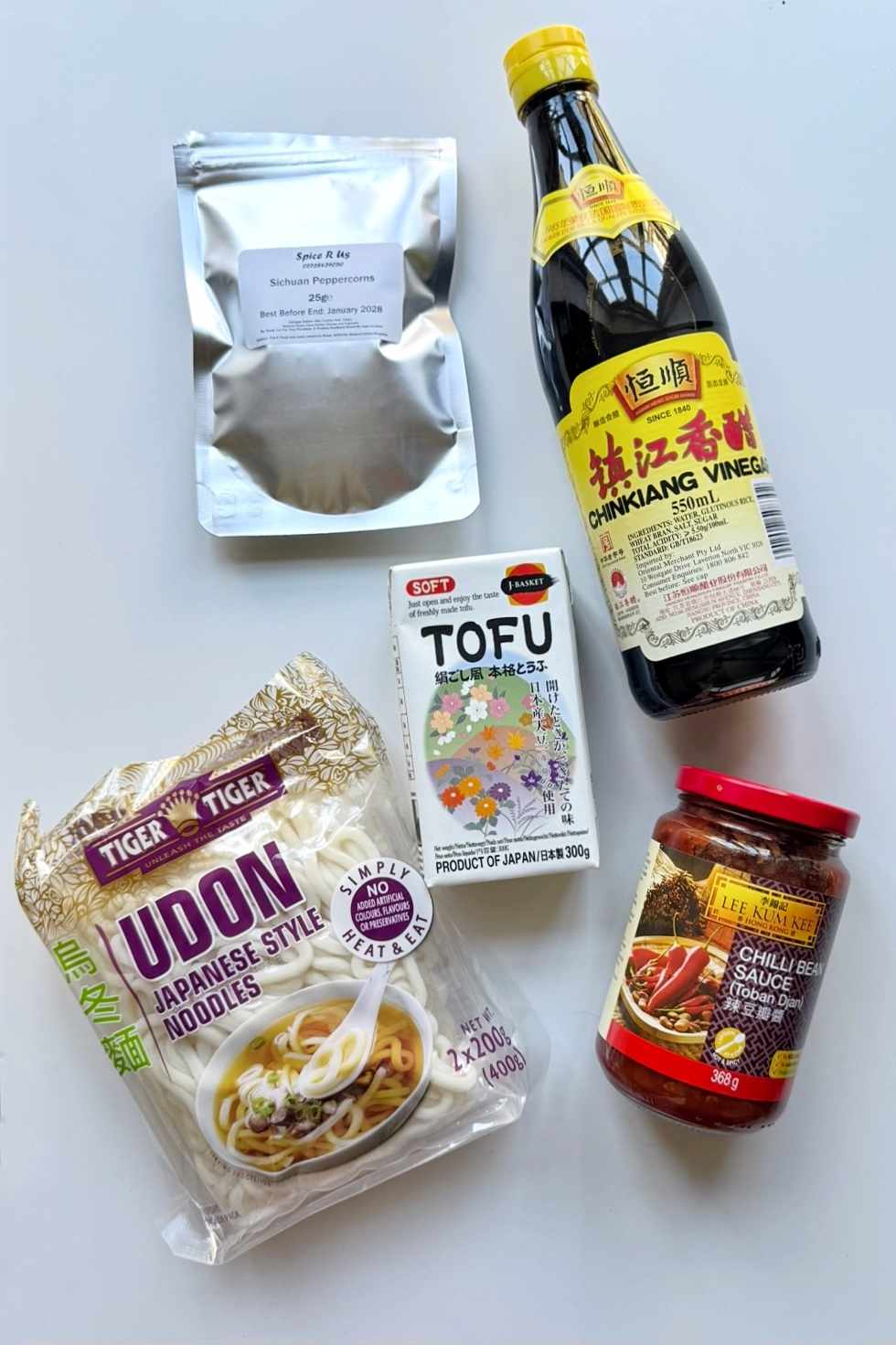 Ingredients for creamy mapo tofu udon laid flat on a white surface, including Tiger udon noodles, soft tofu, Lee Kum Kee chilli bean sauce, Chinkiang vinegar, and Sichuan peppercorns