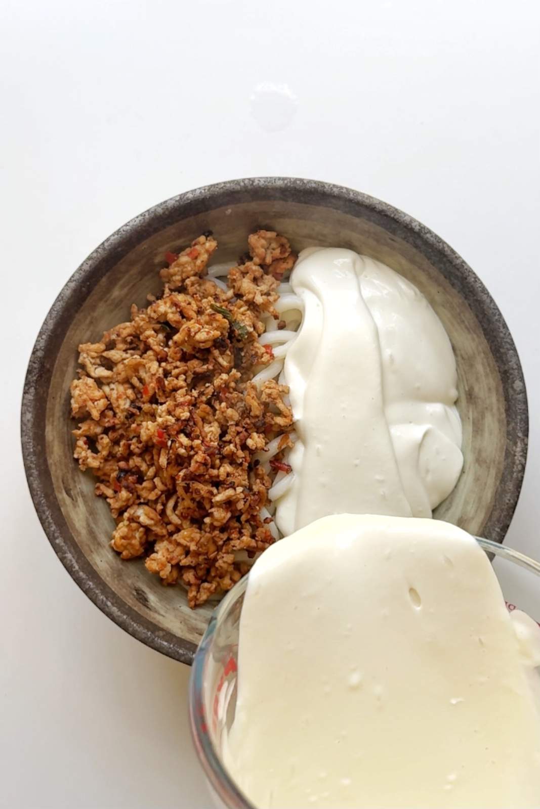 Pouring blended silky tofu cream over cooked udon noodles and spiced mapo meat in a stone bowl