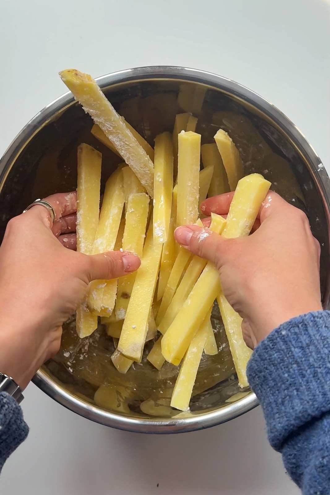 Hands tossing sliced potato batons in a metal bowl with cornstarch coating for nacho fries