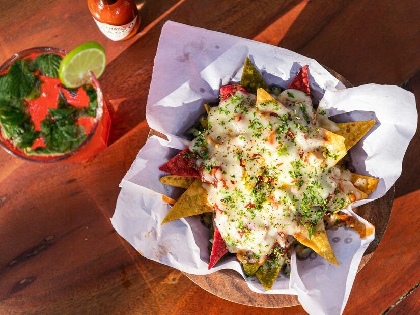 Overhead shot of loaded nachos with melted white cheese, chilli flakes and fresh herbs on multicoloured tortilla chips, served in a parchment-lined wooden bowl with a cocktail alongside