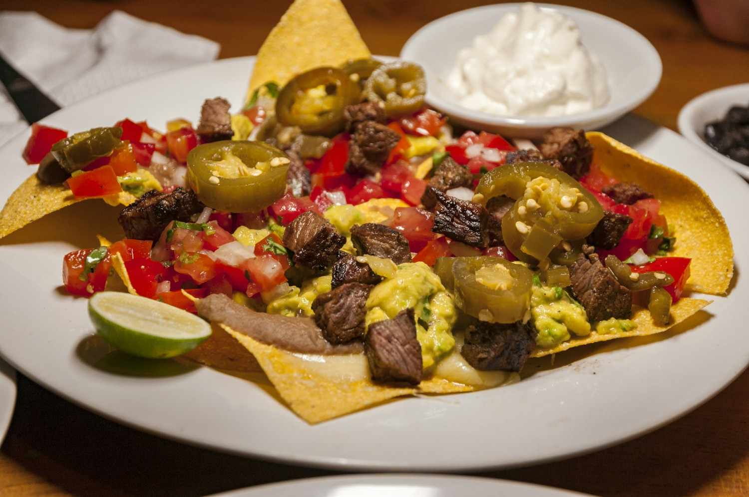 Loaded nachos on a white plate topped with carne asada, pickled jalapeños, pico de gallo, guacamole and a lime wedge, with sour cream and black olives in side bowls