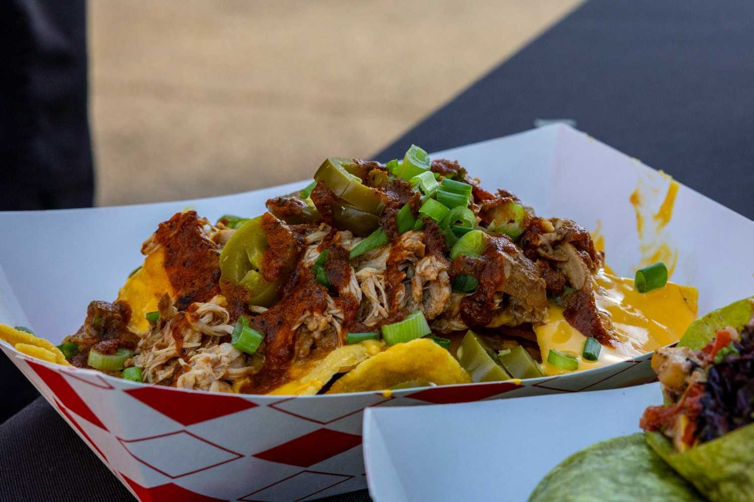 Street food-style loaded nachos in a paper tray with pulled pork, nacho cheese sauce, pickled jalapeños, chilli sauce and sliced spring onions