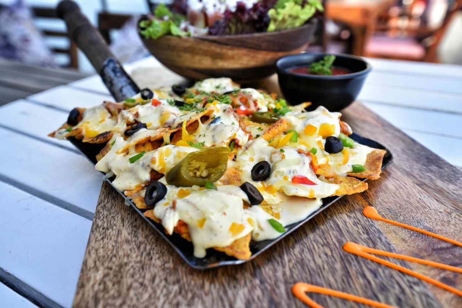 Vegetarian nachos on a black skillet with melted white cheese, pickled jalapeños, black olives, bell peppers, corn and fresh herbs, served with a side of tomato salsa