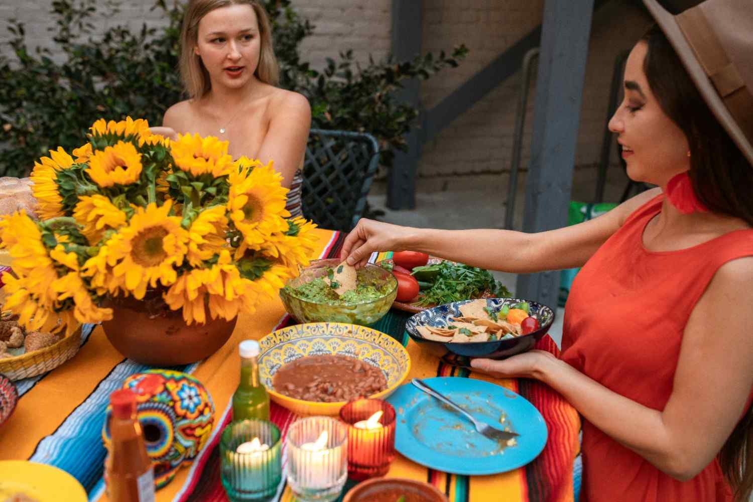 Two women at a colourful outdoor nacho bar spread with guacamole, refried beans, tortilla chips, fresh herbs and hot sauce on a striped table runner