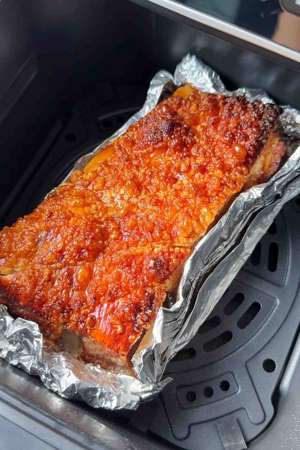 Crispy pork belly with golden, crackling skin resting in an air fryer basket lined with foil.