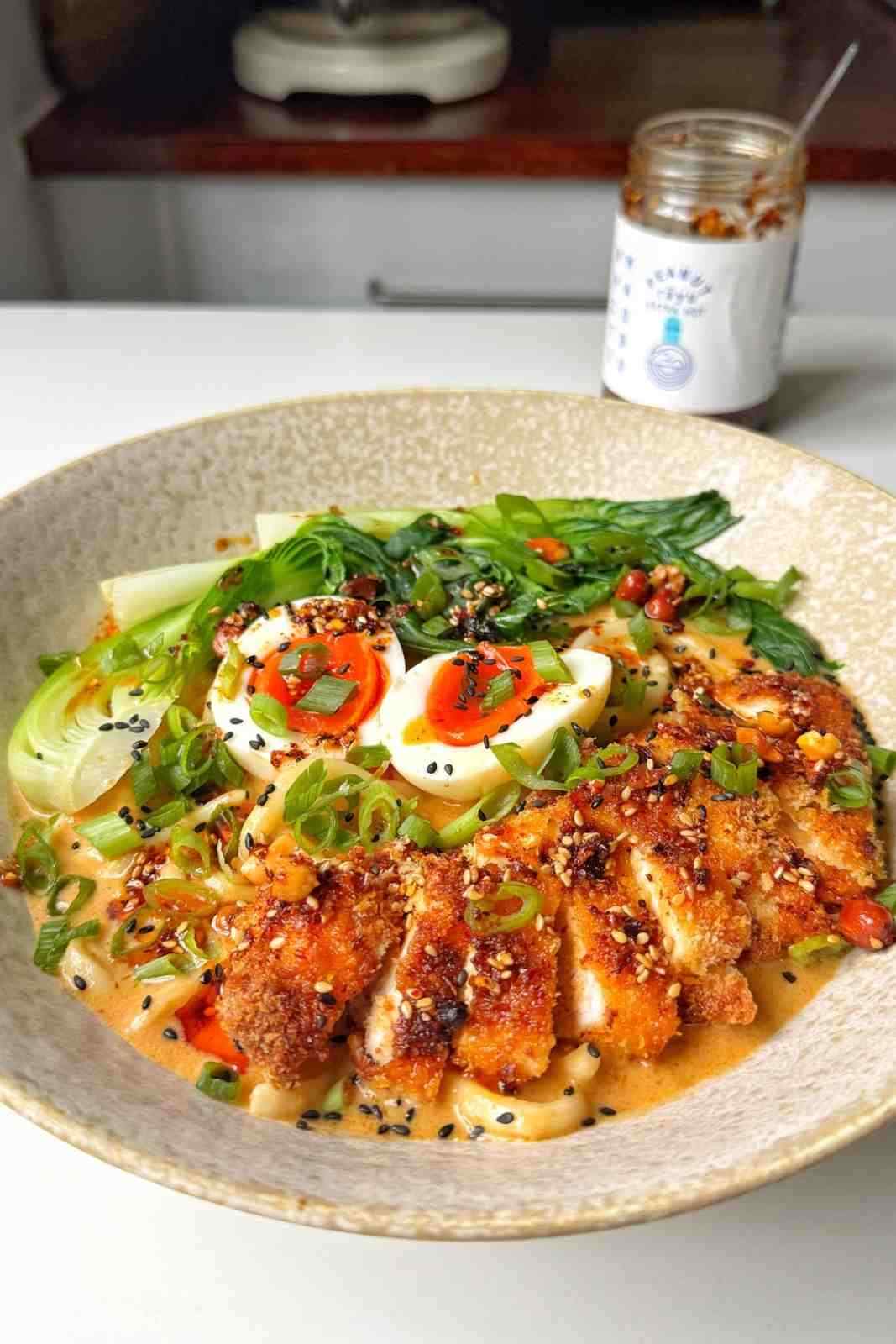 A vibrant ramen bowl topped with crispy air-fried chicken katsu, soft-boiled jammy eggs, bok choy, and chili oil.