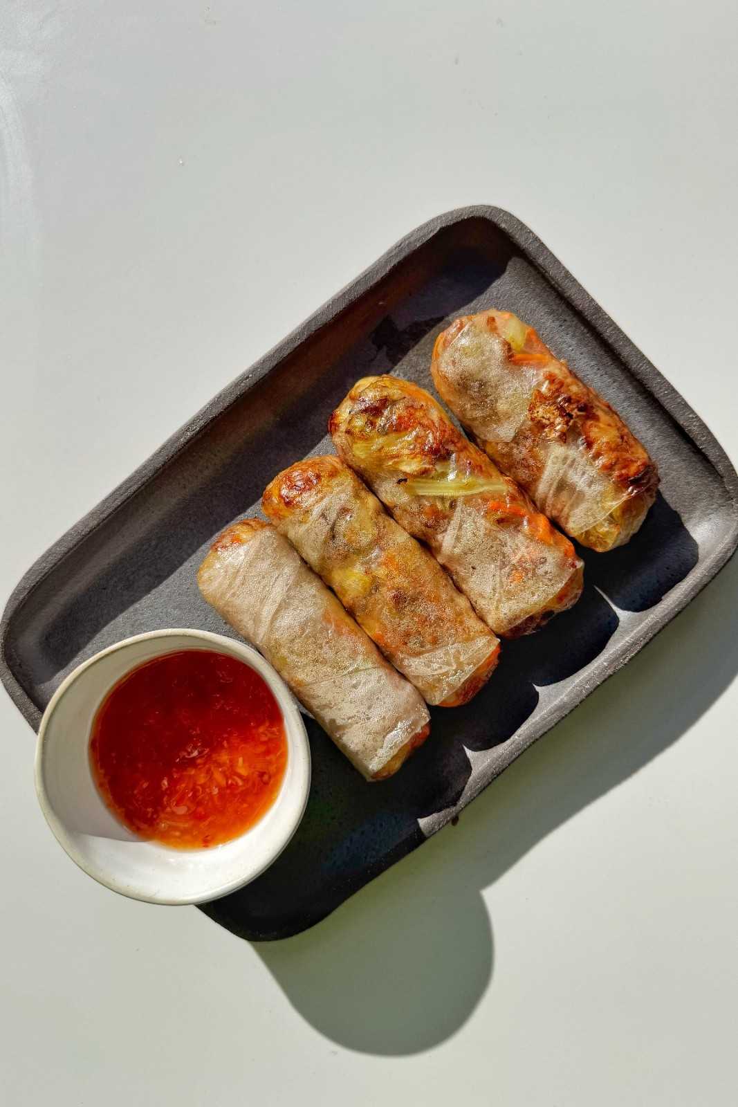 Four crispy air-fried spring rolls on a dark ceramic tray served with a side of sweet chili dipping sauce.