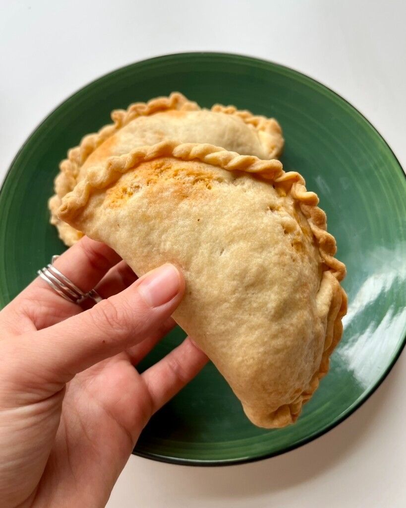 A hand holding a golden-brown air-fried empanada with a braided crust, served on a green plate.