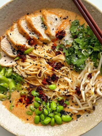 A bowl of creamy ramen noodles topped with sliced chicken, cilantro, bean sprouts, edamame, scallions, chili oil, and black sesame seeds with chopsticks resting on the side.