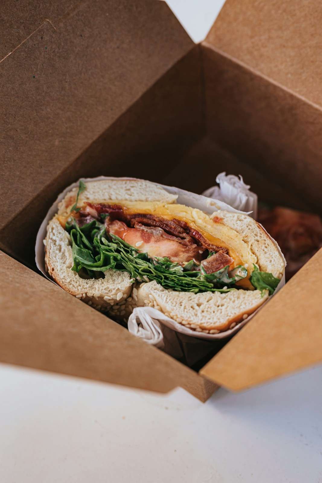 BLT bagel sandwich with crispy bacon, fresh greens, and tomato on a sesame bagel in a takeout box