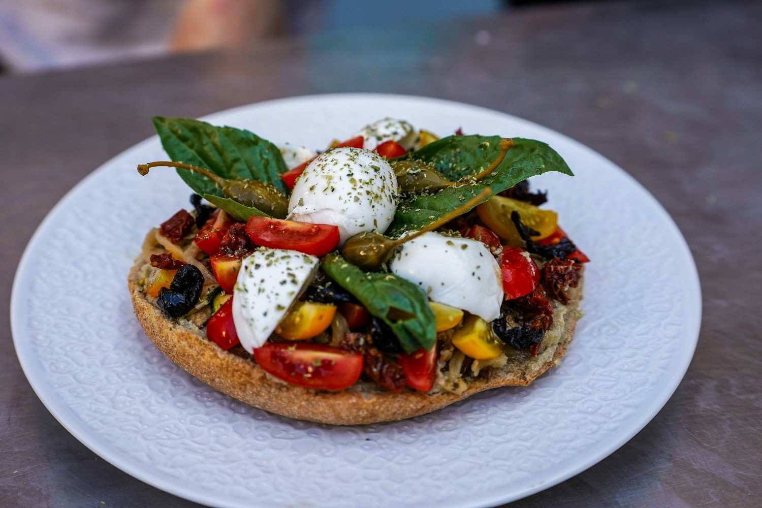 Open-faced caprese bagel with fresh mozzarella, cherry tomatoes, sun-dried tomatoes, pesto, and basil leaves on a white plate