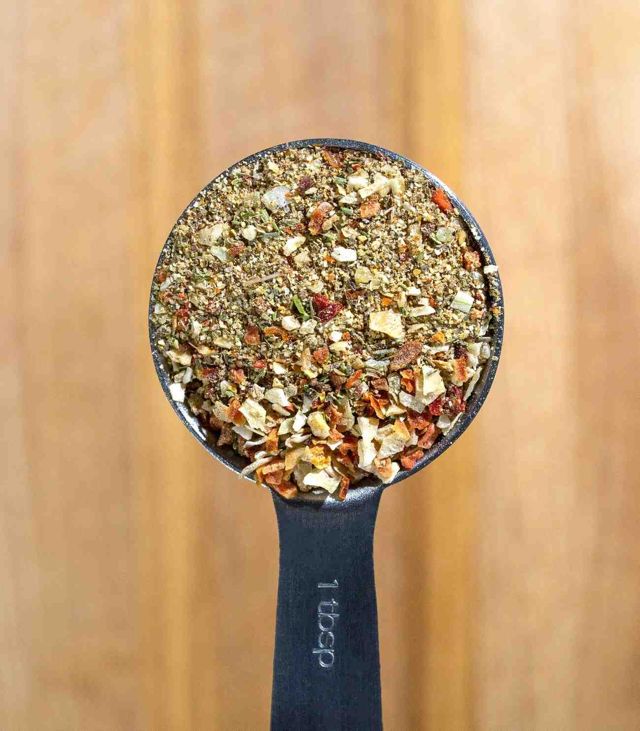 Close-up of a tablespoon filled with Montreal steak seasoning showing coarse black pepper, garlic, red pepper flakes, and herbs