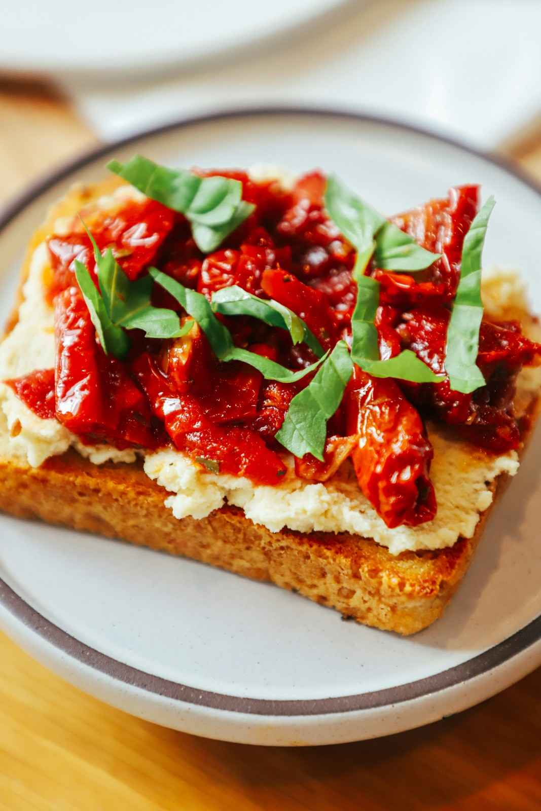 Toasted bread topped with cream cheese, sun-dried tomatoes, and fresh basil on a white plate
