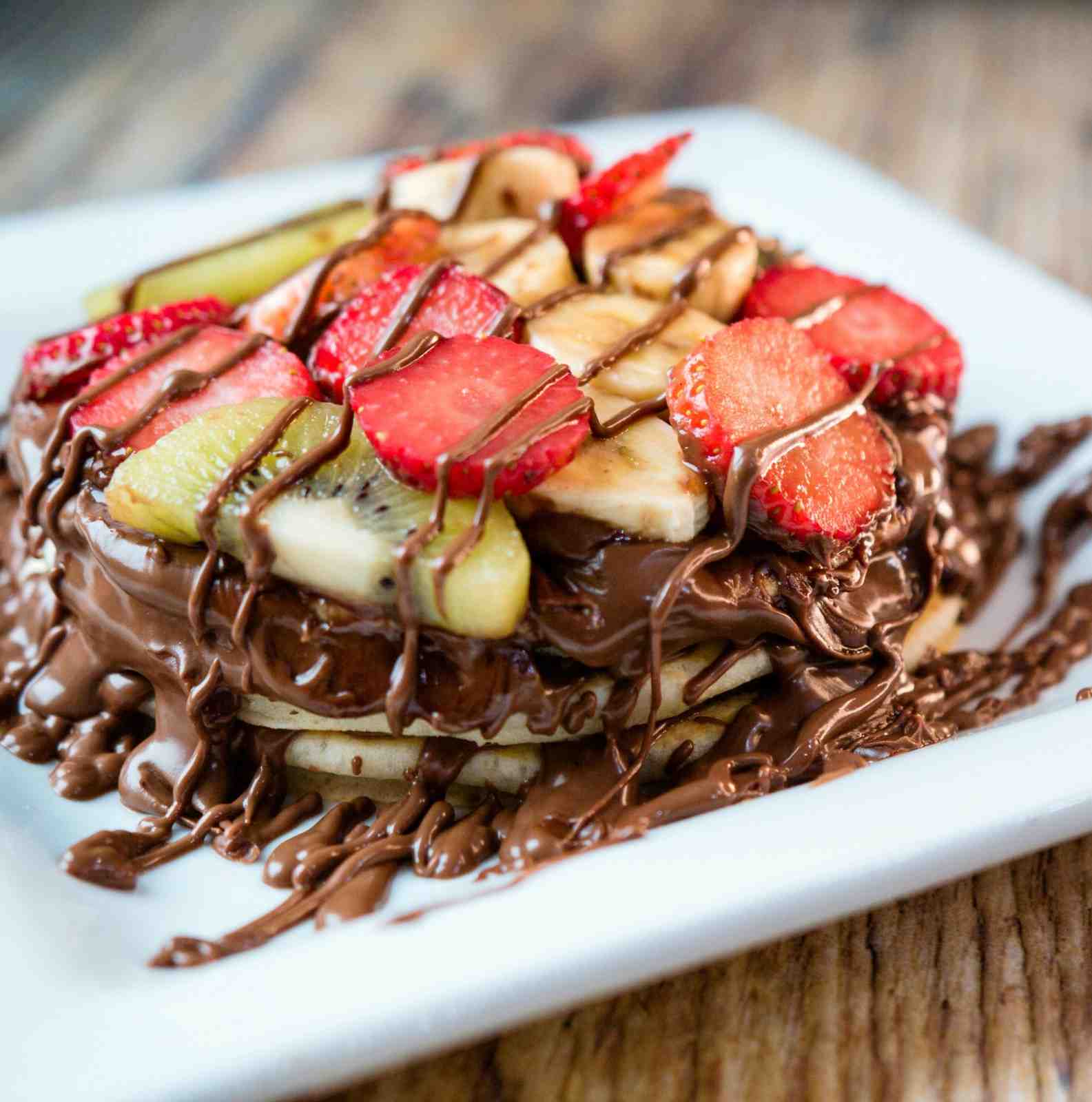 Stack of Nutella-drizzled pancakes topped with fresh strawberries, sliced banana, and kiwi on a white plate