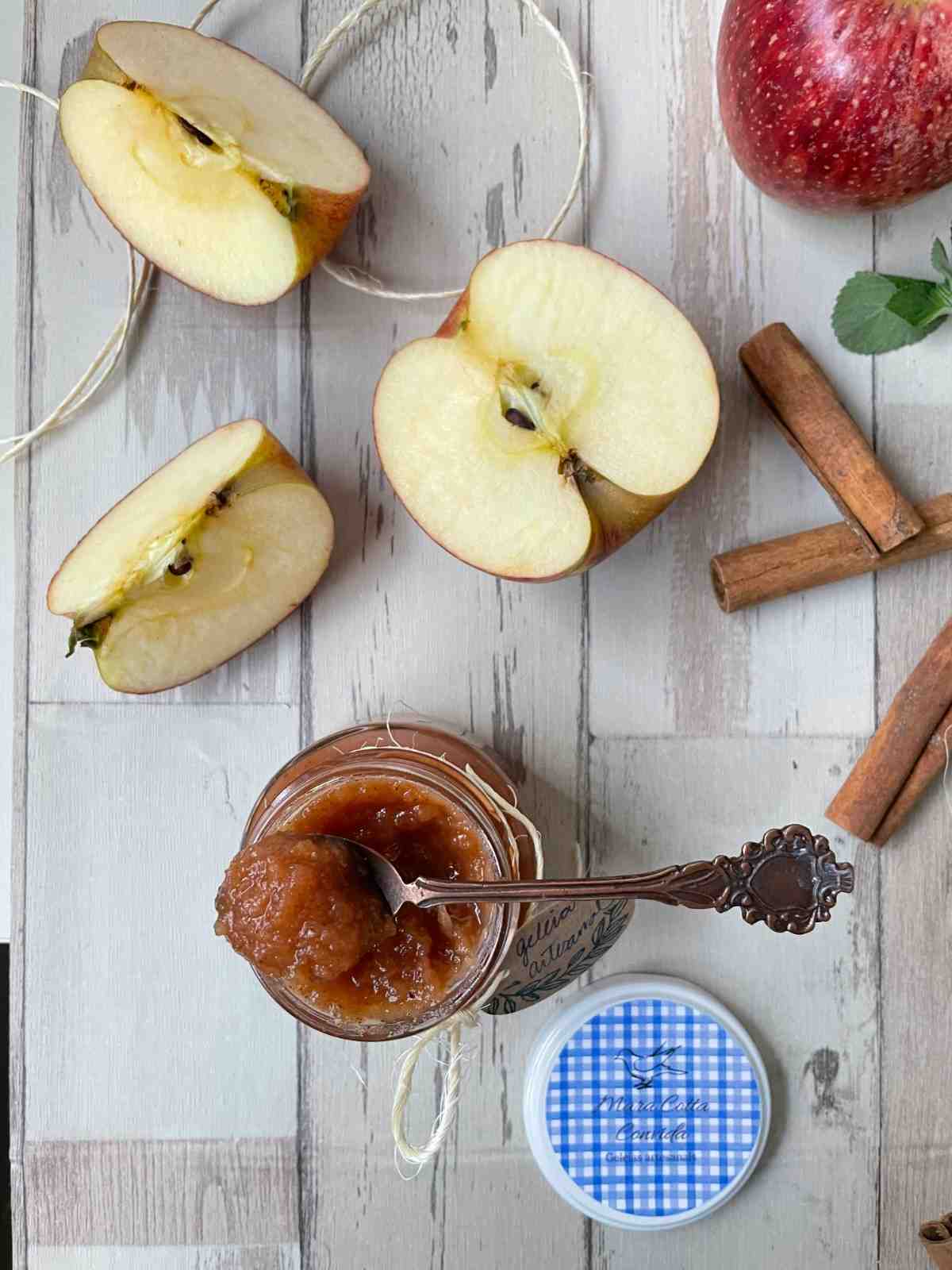 Jar of homemade apple butter with a spoon surrounded by halved apples and cinnamon sticks on a rustic white wooden surface