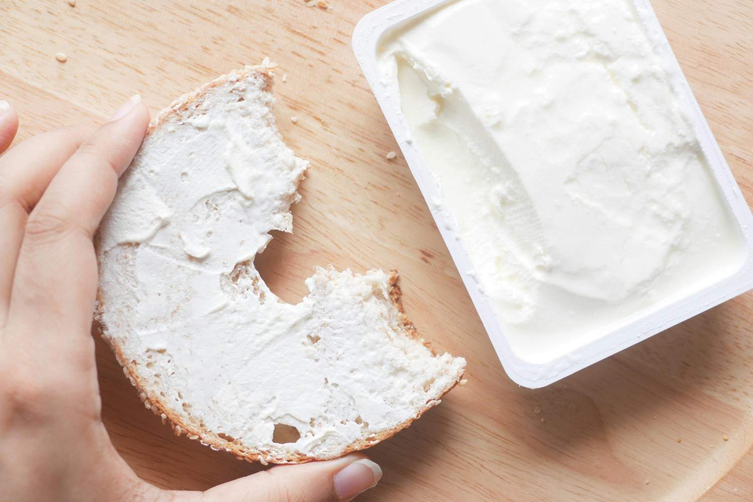 Hand holding a bitten bagel half spread with cream cheese next to an open tub of cream cheese on a wooden board