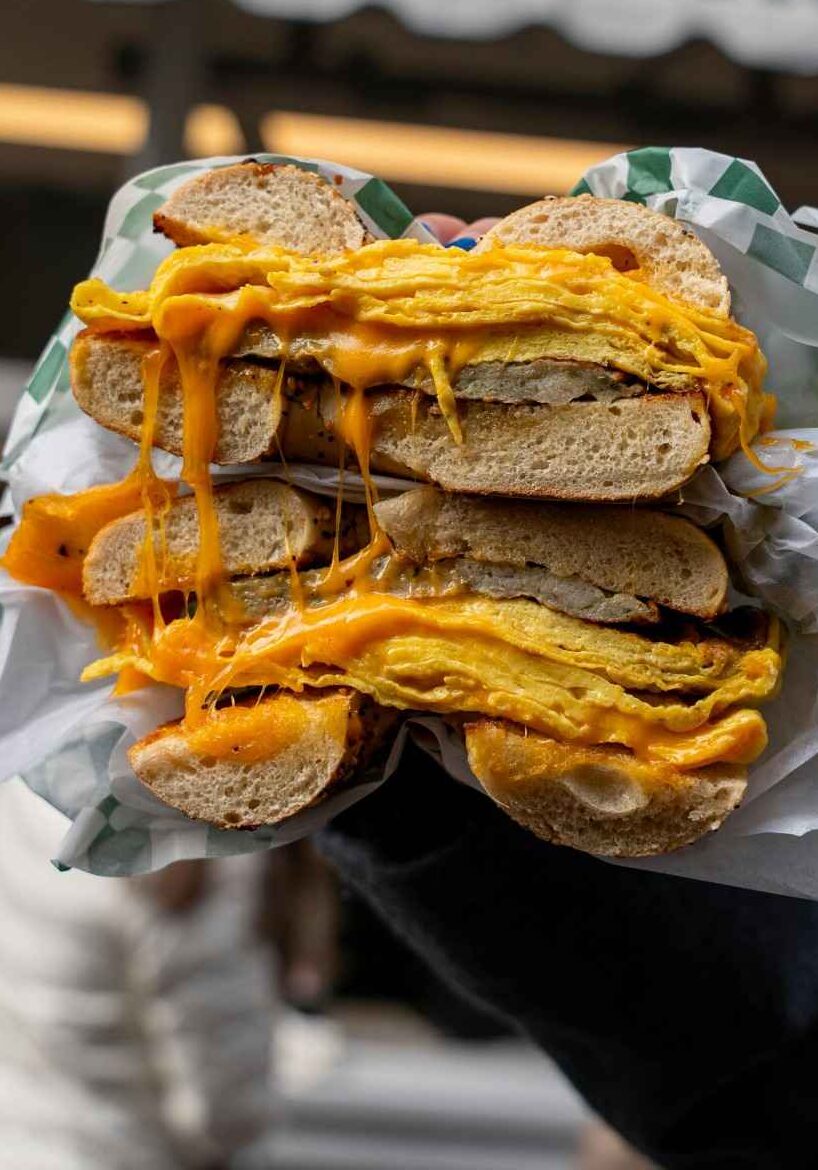 Bacon egg and cheese bagel with melted American cheese dripping down the sides, held up outside a bagel shop