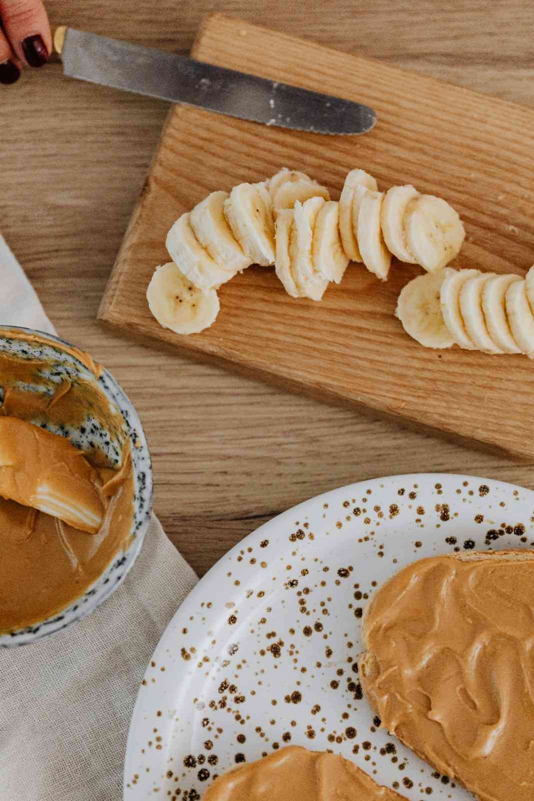 Peanut butter spread on toast with sliced banana and a bowl of peanut butter on a wooden board