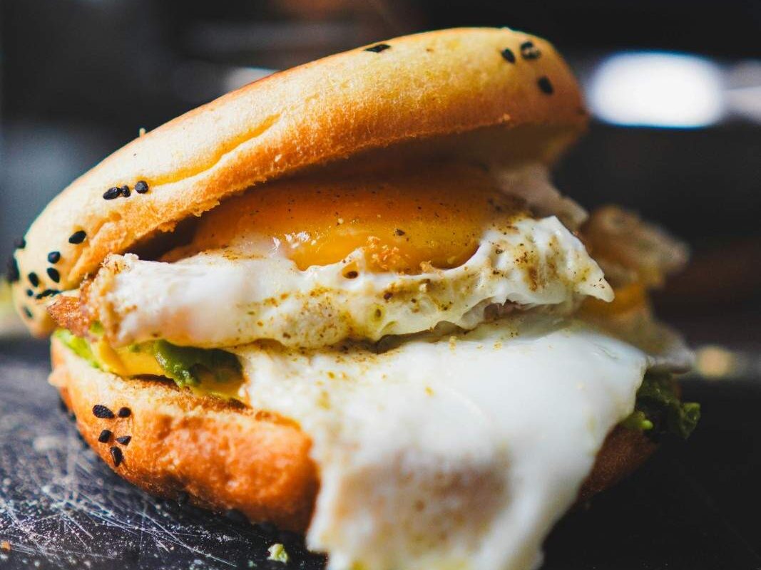 Sesame bagel filled with mashed avocado and a fried egg with a runny yolk on a dark surface