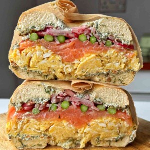 Stacked smoked salmon bagel halves showing layers of the best bagel toppings including dill cream cheese, scrambled eggs, pickled red onions, and asparagus on a wooden cutting board