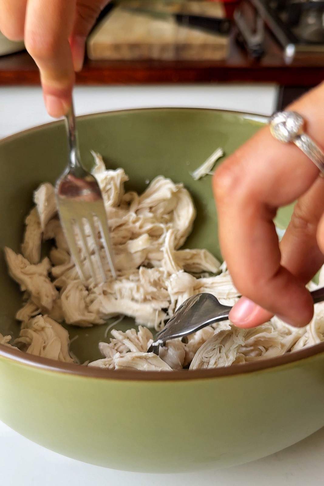 Hands shredding rotisserie chicken with two forks in a green bowl, showing the easy prep method for a baked potato bar protein topping.