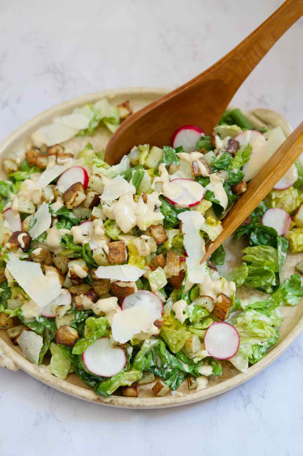 A fresh Caesar salad with chopped romaine, crispy croutons, shaved parmesan, sliced radishes, and creamy Caesar dressing in a serving bowl with wooden tongs — the viral TikTok baked potato topping.