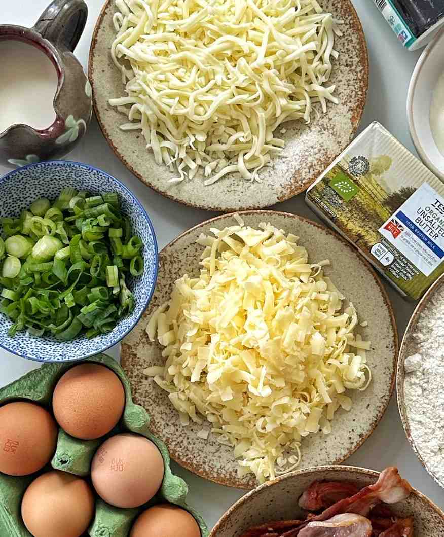 A flatlay of baked potato bar essentials including two plates of freshly shredded cheddar and mozzarella cheese, sliced scallions, unsalted butter, eggs, and bacon.