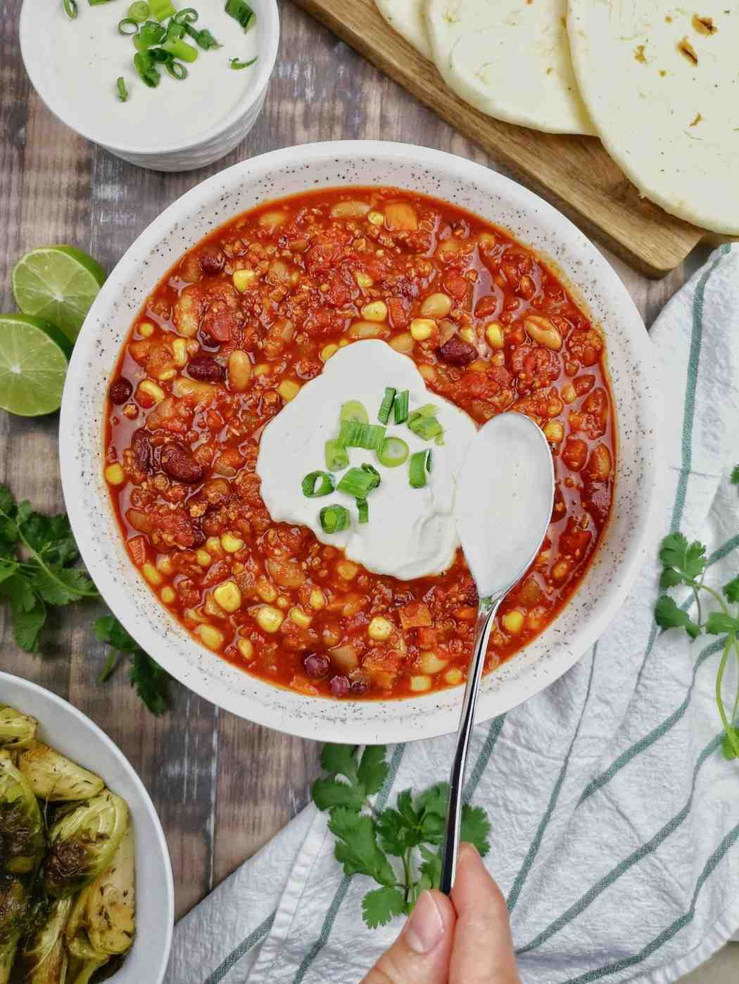 A bowl of hearty beef chili with beans and corn topped with a dollop of sour cream and chopped scallions, served alongside lime wedges and fresh cilantro.