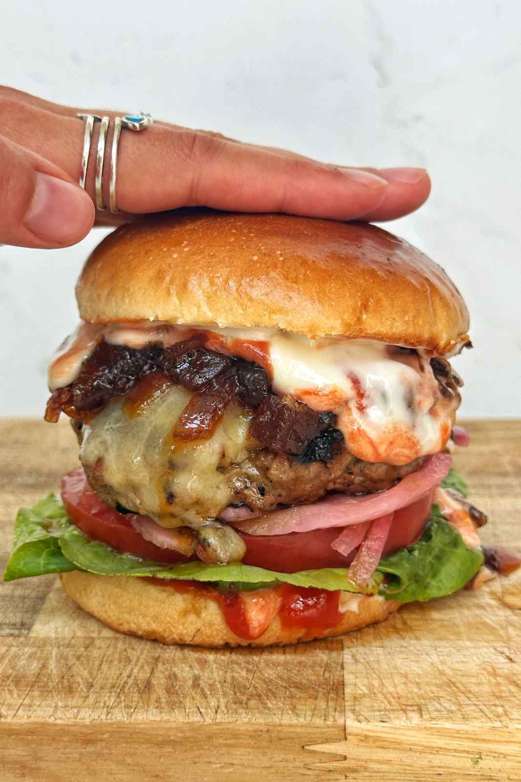 Close-up of a homemade cheeseburger on a toasted brioche bun with melted cheese, crispy bacon, caramelized onions, pickled red onion, tomato, lettuce, and burger sauce dripping down the sides.