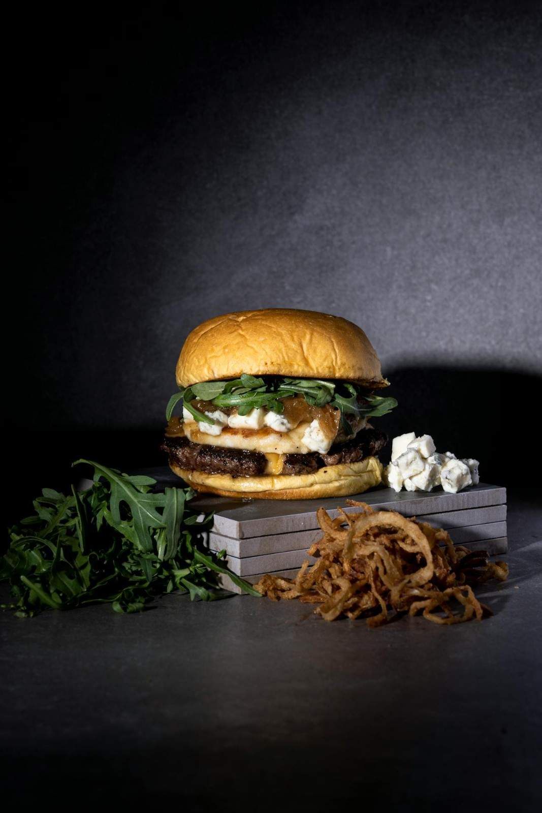 A gourmet burger topped with blue cheese crumbles, caramelized onions, and fresh arugula on a toasted brioche bun, with a pile of crispy fried onions and extra blue cheese on the side.