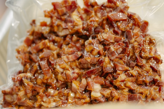 A close-up of finely chopped bacon glistening with rendered fat and dark, sticky glaze, ready to be cooked down into bacon jam.