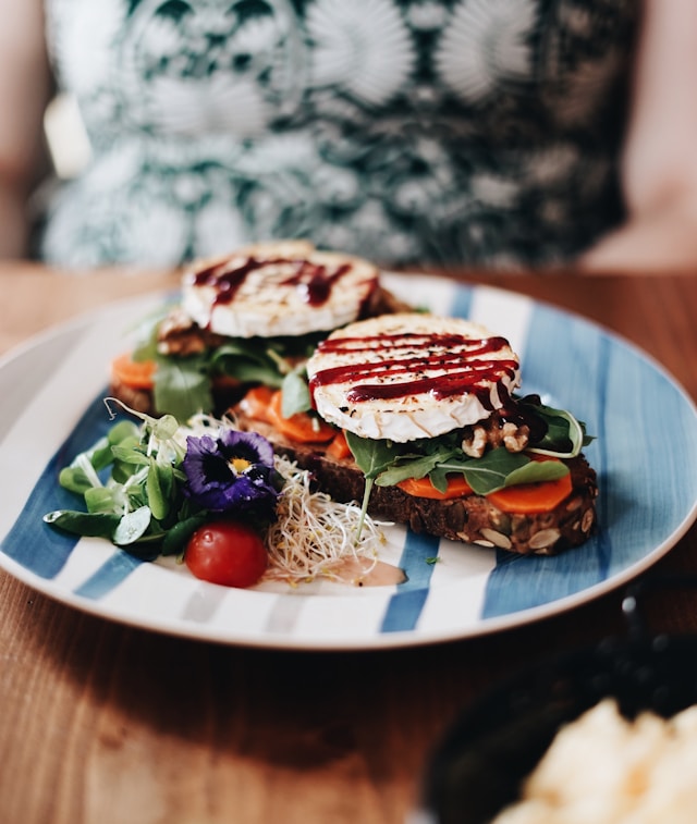 An open-faced sandwich topped with thick slices of grilled goat cheese, balsamic glaze, arugula, and tomato on dark seeded bread.