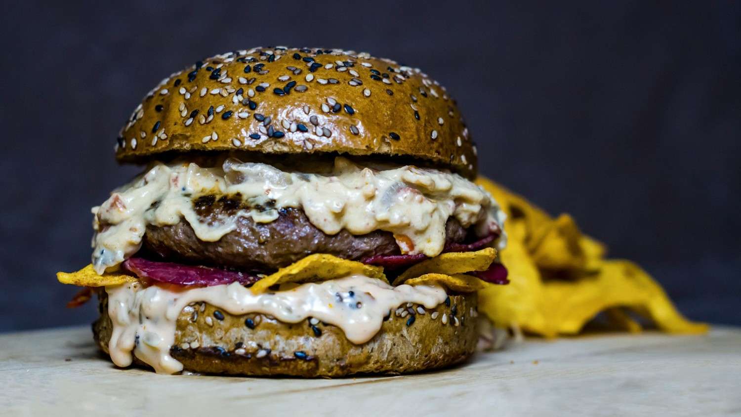 A juicy beef burger on a sesame seed bun topped with creamy chunky pimento cheese spread, pickled red onion, and crispy tortilla chips on the side.