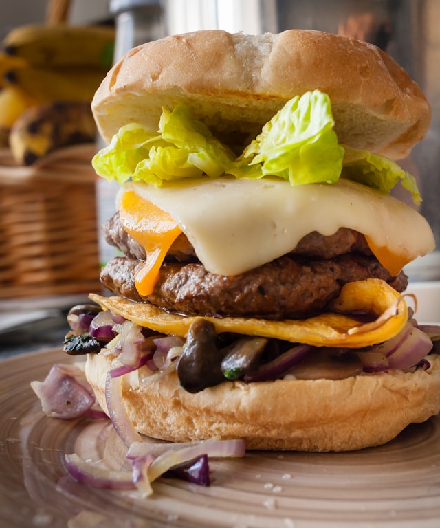 A tall double-patty burger topped with melted smoked gouda and cheddar, butter lettuce, caramelized onions, and sautéed mushrooms on a toasted bun.