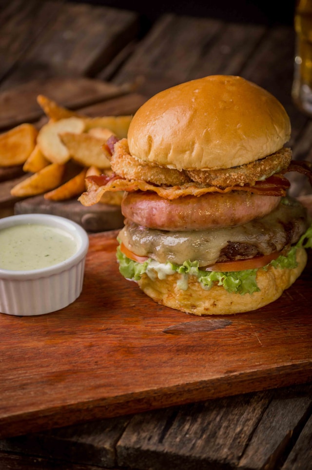 A stacked gourmet burger with crispy fried pickle chips, a pork patty on top of a beef patty, melted cheese, bacon, lettuce, and tomato on a brioche bun, served with potato wedges and a ramekin of dipping sauce.