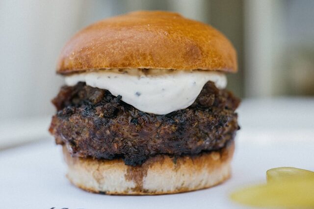 A small gourmet slider on a brioche bun topped with a thick dollop of creamy garlic aioli, served on a white restaurant plate with a pickle on the side.