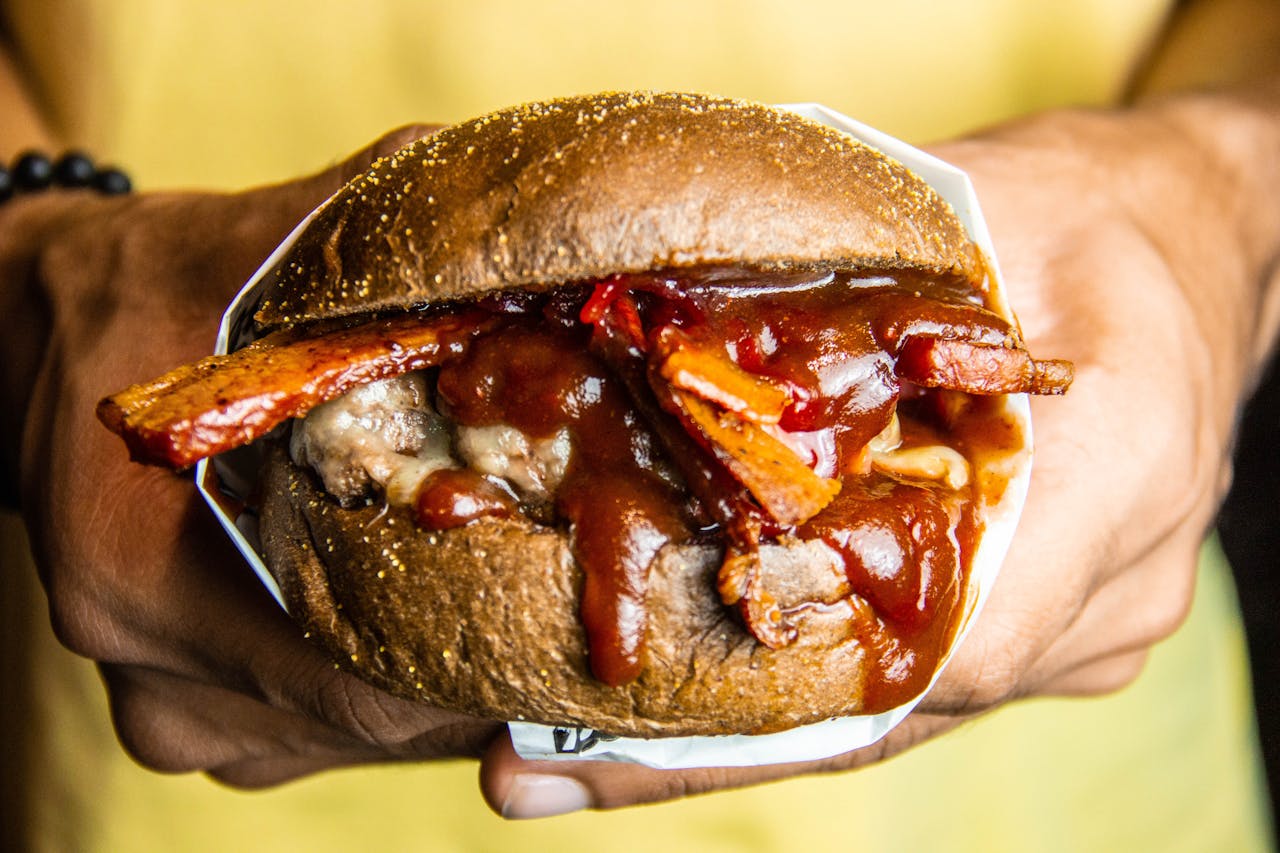 Hands holding a burger smothered in dripping BBQ sauce with crispy bacon and melted cheese spilling out from the sides of a sesame bun.