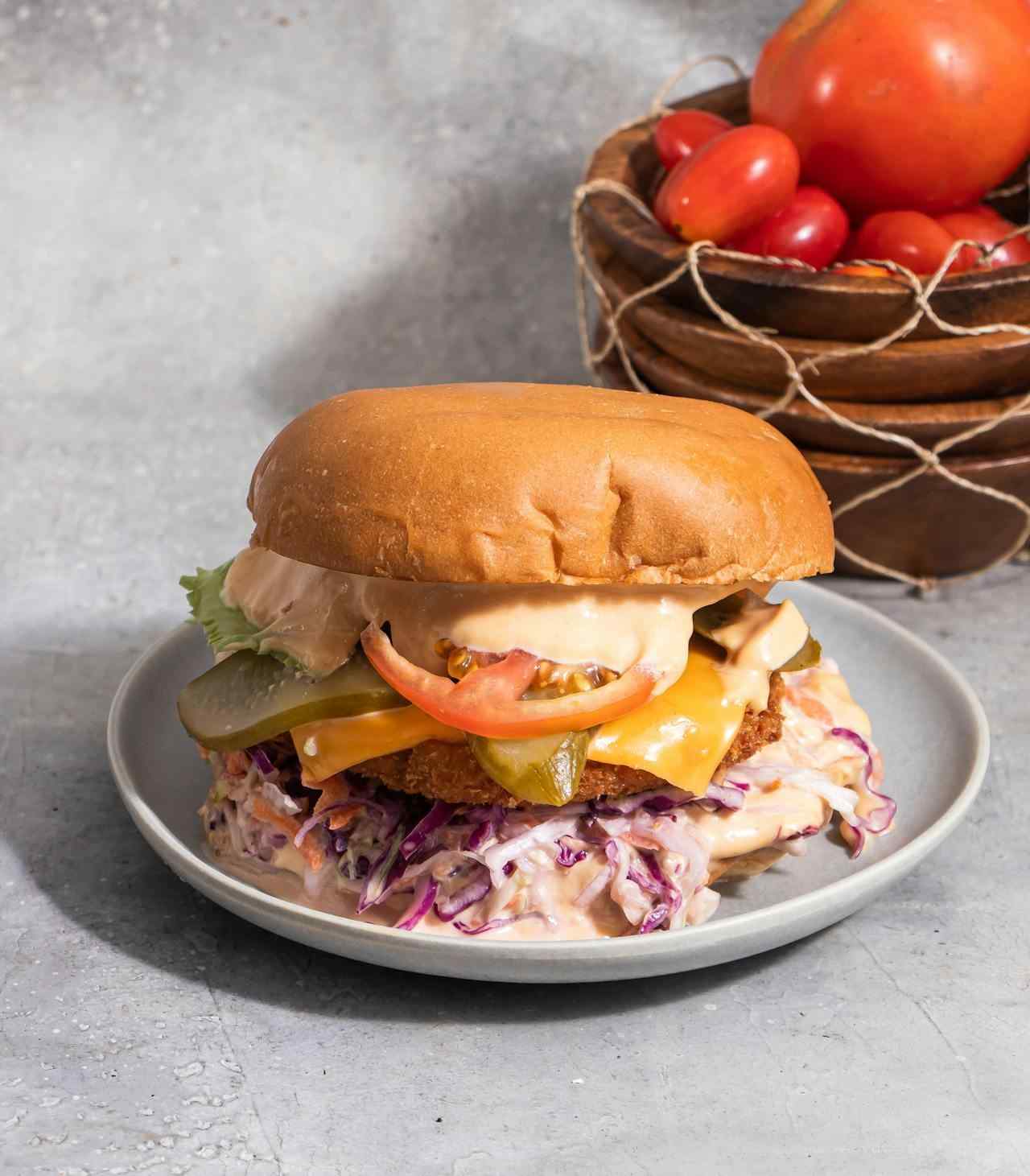 A crispy breaded chicken burger on a soft bun topped with creamy pink thousand island dressing, a slice of cheddar, a pickle, tomato, and lettuce, sitting on a bed of coleslaw with a bowl of fresh tomatoes in the background.